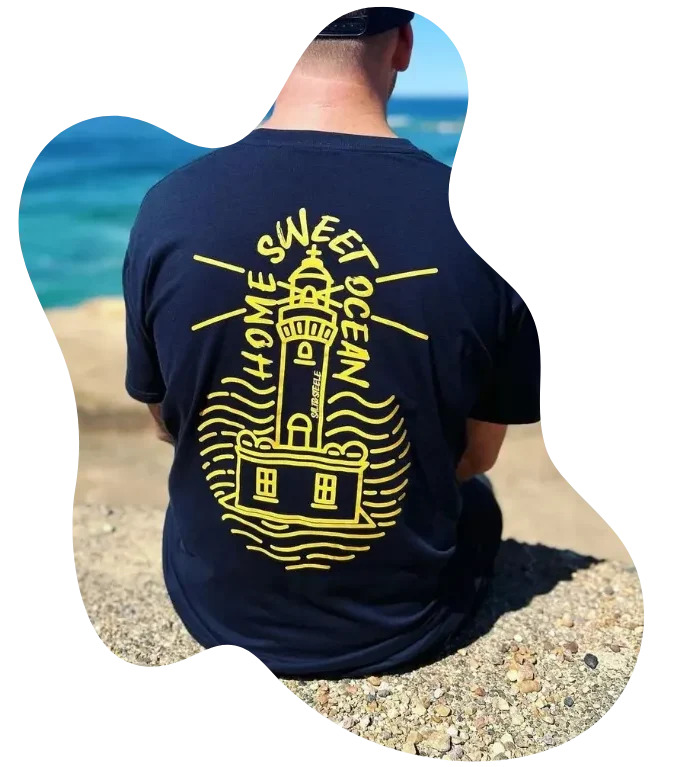 Back view of a customer sitting by the beach, wearing a navy T-shirt with a yellow lighthouse design from the “Wear the Roots – Grow the Future” collection